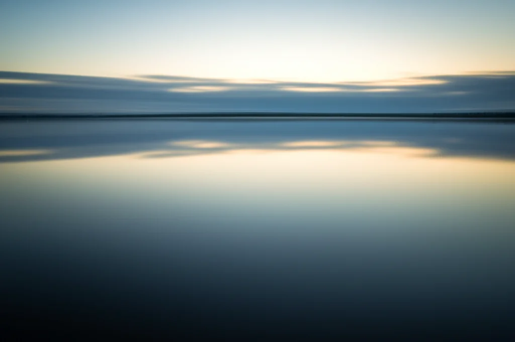 A wide-angle landscape shot of a calm lake or river at dawn, with soft light reflecting on the water. Use a 10mm wide-angle lens with a long exposure time to create smooth water and clouds, evoking a sense of quiet focus or preparation.