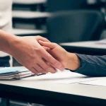 Immagine simbolica del contract cheating: una mano che passa di nascosto un compito universitario a un'altra mano sotto un banco, in un'aula universitaria. Illuminazione soffusa, obiettivo 50mm, focus selettivo sulle mani, stile documentaristico.